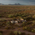 Aerial view of a roofless white-walled adobe ruin among desert mesquite, a mountain range in the hazy distance.