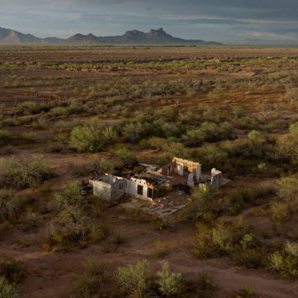 Aerial view of a roofless white-walled adobe ruin among desert mesquite, a mountain range in the hazy distance.