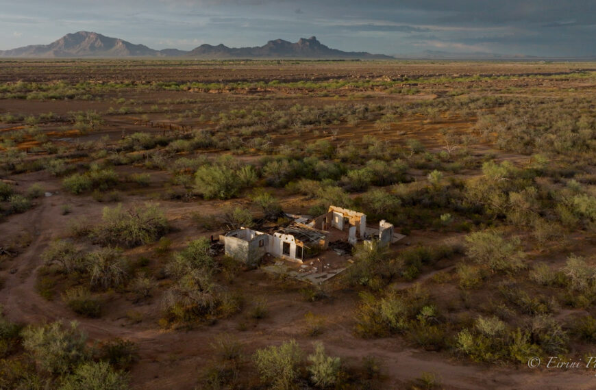 Aerial view of a roofless white-walled adobe ruin among desert mesquite, a mountain range in the hazy distance.