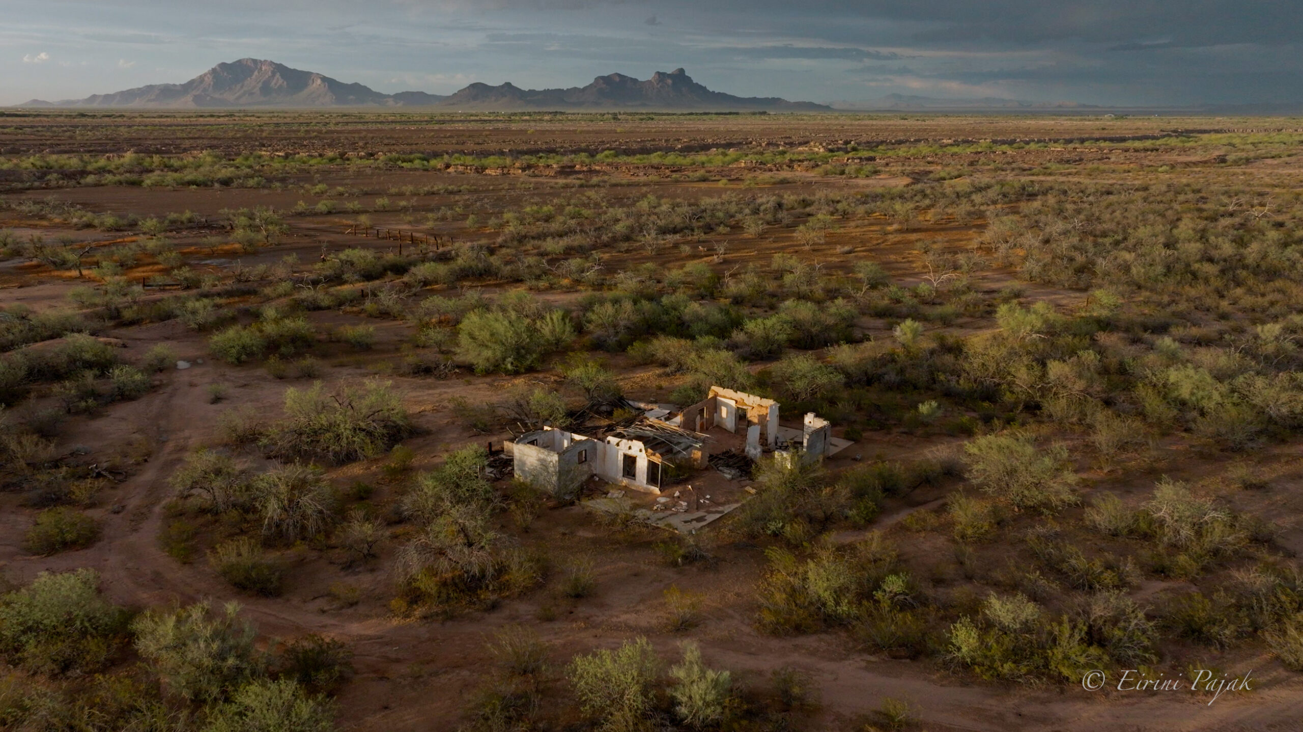 Aerial view of a roofless white-walled adobe ruin among desert mesquite, a mountain range in the hazy distance.