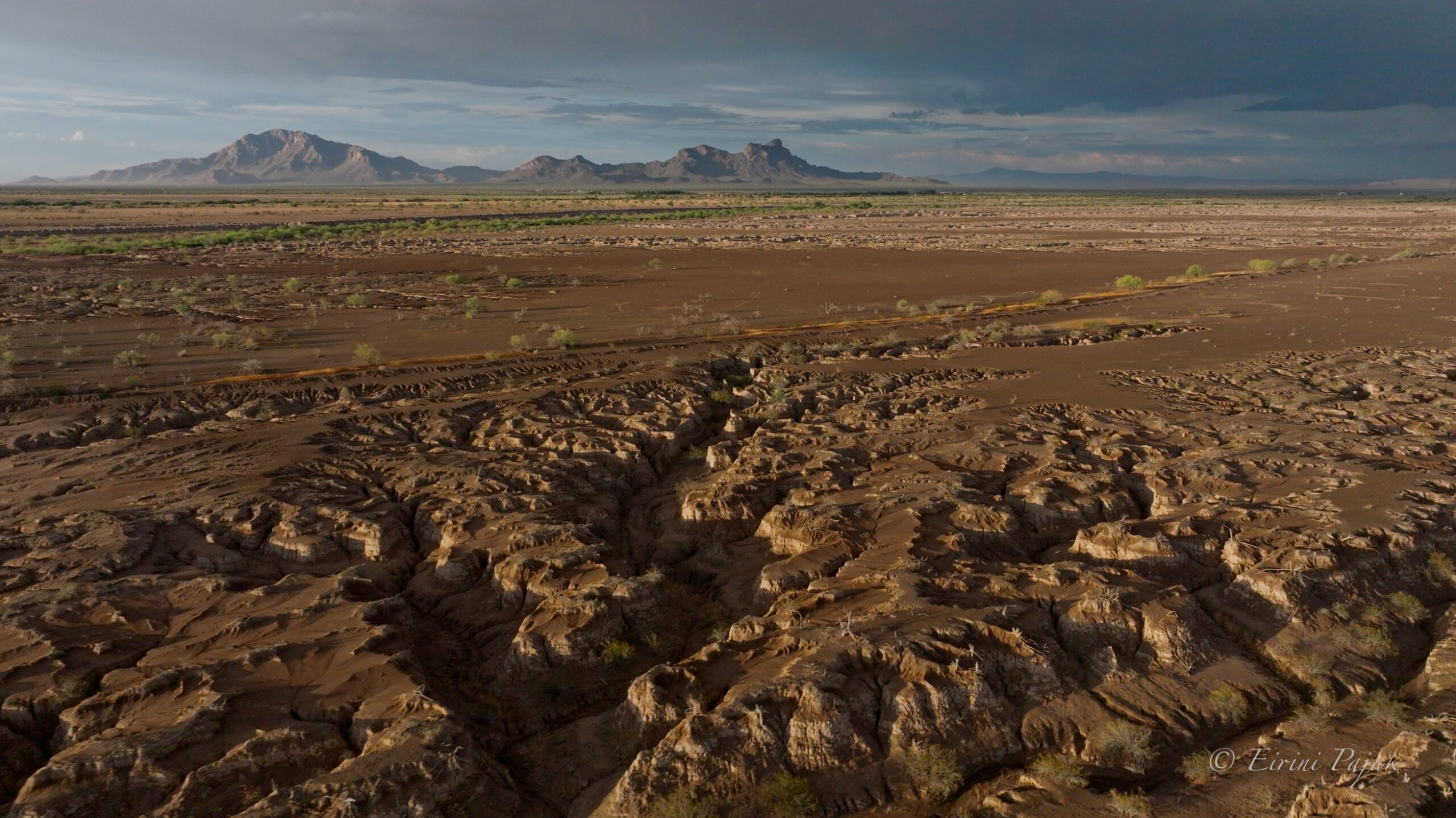 Aerial view of deeply eroded, fan-shaped channels in bare desert soil, mountains and a dark storm sky beyond.