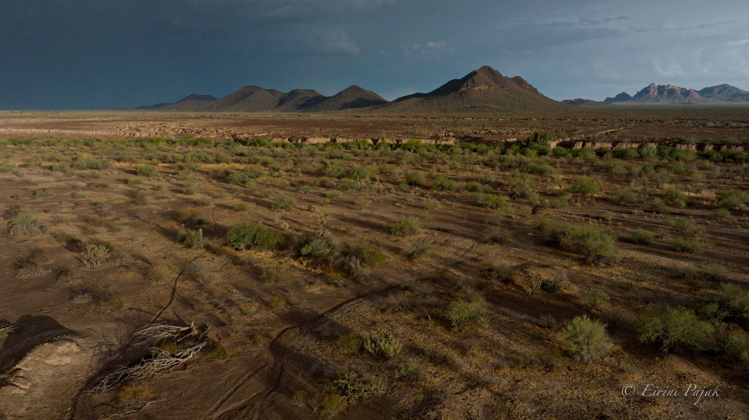 Aerial view of desert brush and a sunken wash leading toward dark conical peaks beneath a heavy storm sky.