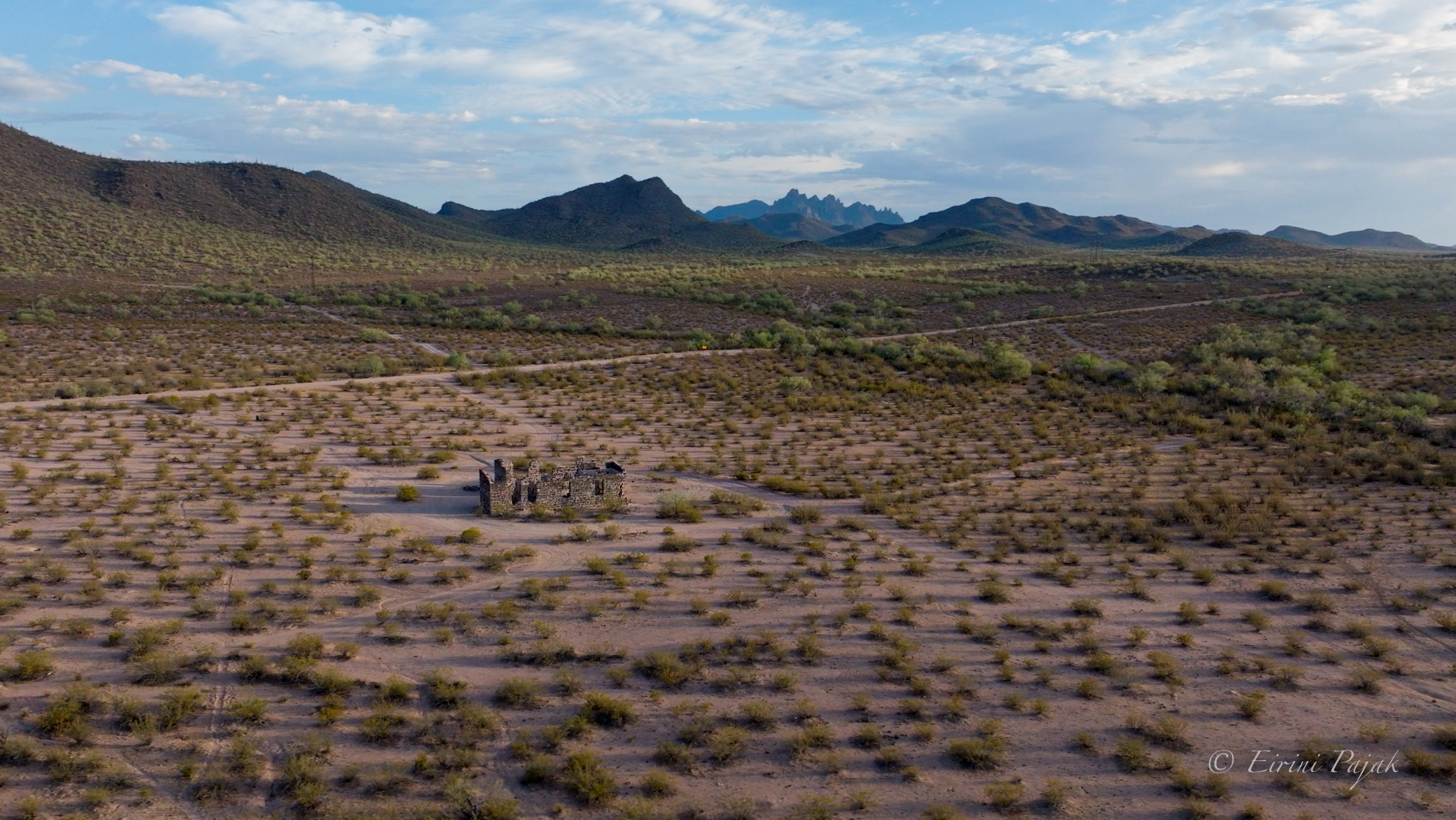 Aerial view of graffiti-marked concrete smelter foundations among green desert brush, with low mountains and a dirt road behind.