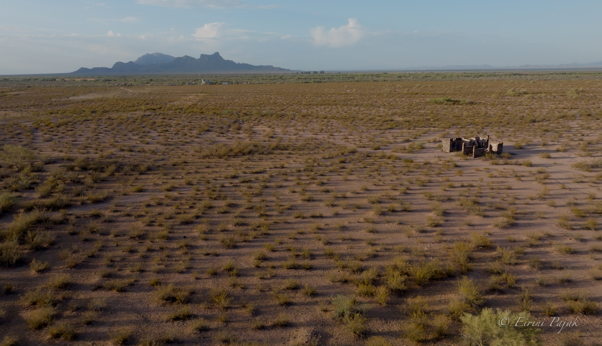Aerial view of a small stone ruin on flat, sparsely vegetated desert, a low mountain range on the far horizon.