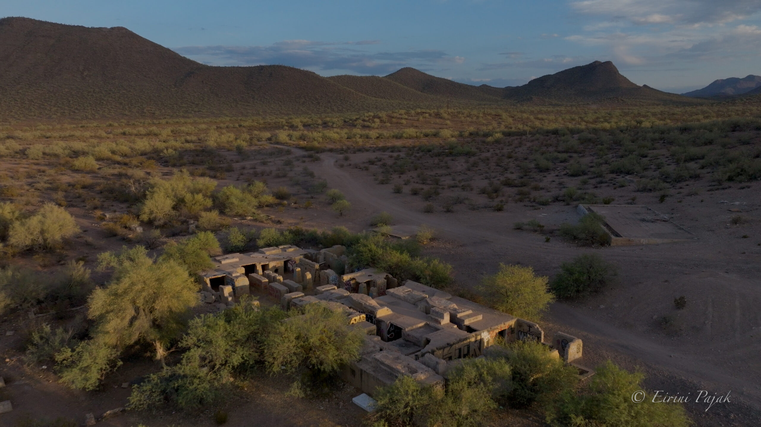 A line of freestanding concrete walls and pillars runs across a desert wash, backed by rolling hills and scattered saguaro.