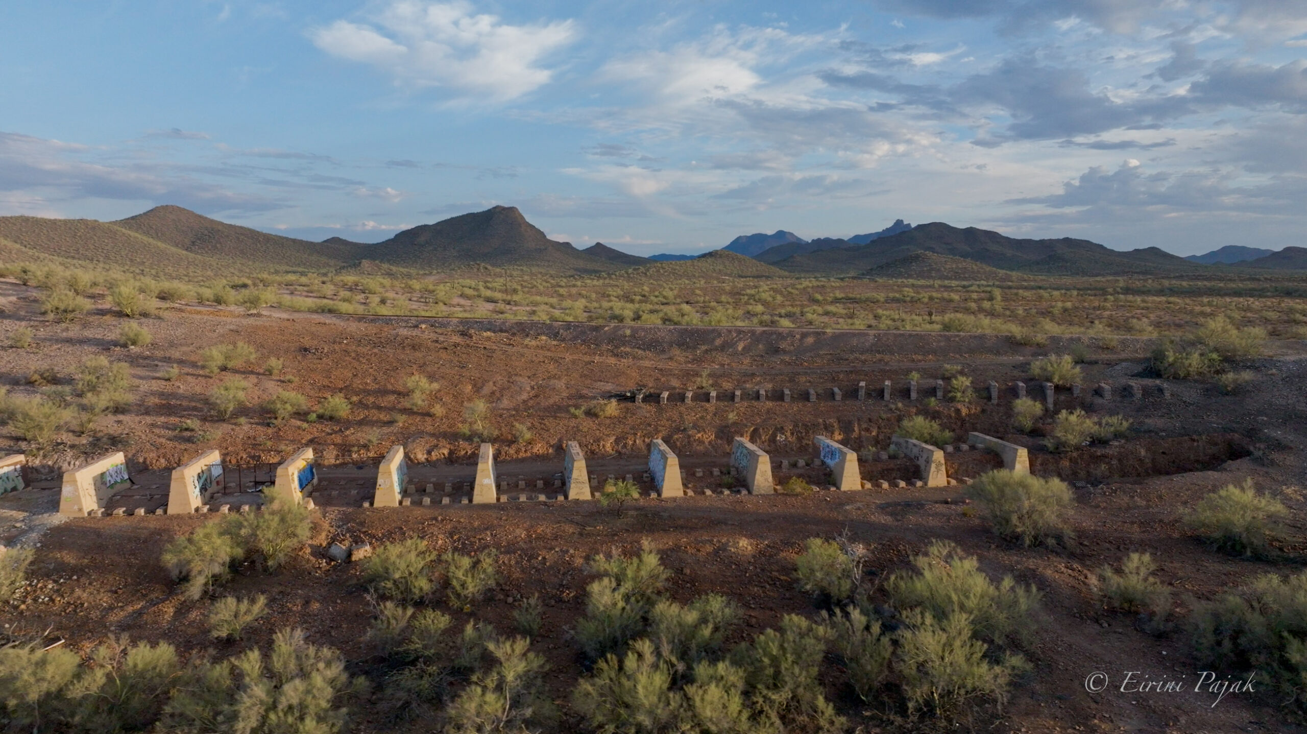 A line of freestanding concrete walls and pillars runs across a desert wash, backed by rolling hills and scattered saguaro.