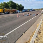Portable weigh scales sit in a traffic lane bordered by orange cones. A yellow Loves fuel tanker, other commercial trucks, and law enforcement vehicles are visible on the roadway in the background. A white inspection trailer is parked on the right shoulder.