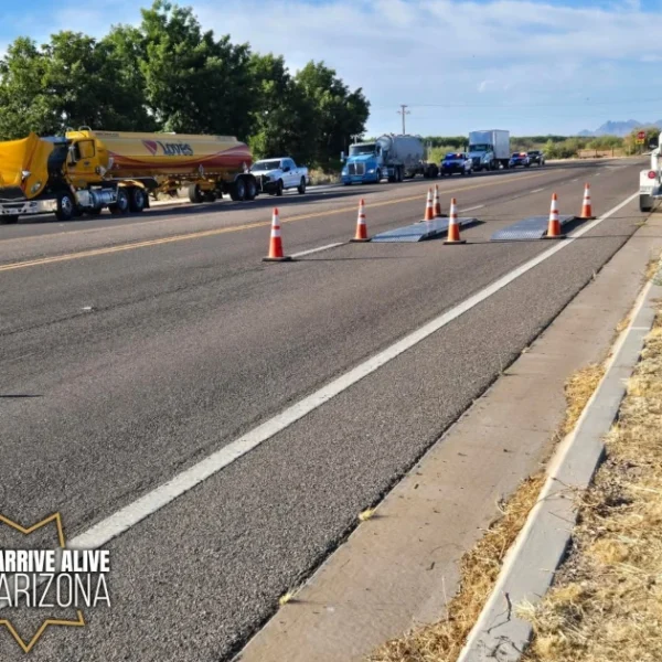 Portable weigh scales sit in a traffic lane bordered by orange cones. A yellow Loves fuel tanker, other commercial trucks, and law enforcement vehicles are visible on the roadway in the background. A white inspection trailer is parked on the right shoulder.