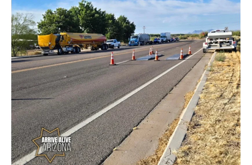 Portable weigh scales sit in a traffic lane bordered by orange cones. A yellow Loves fuel tanker, other commercial trucks, and law enforcement vehicles are visible on the roadway in the background. A white inspection trailer is parked on the right shoulder.