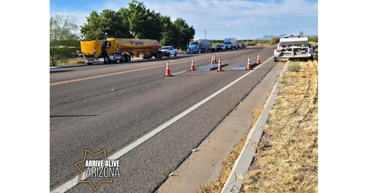 Portable weigh scales sit in a traffic lane bordered by orange cones. A yellow Loves fuel tanker, other commercial trucks, and law enforcement vehicles are visible on the roadway in the background. A white inspection trailer is parked on the right shoulder.