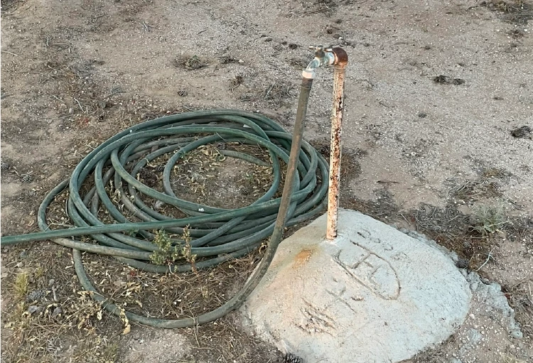 A coiled green hose lying on dirt next to a rusty metal pipe topped with a spigot mounted on a small concrete base with markings at Florence Memorial Park