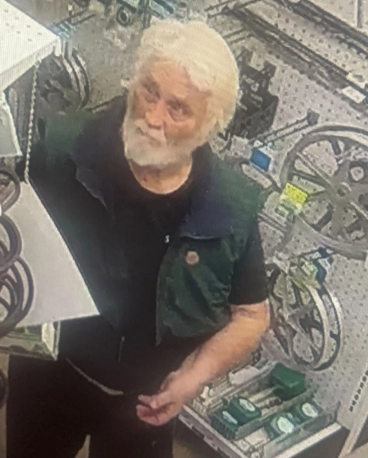 Surveillance image of an older man with white hair and a full white beard wearing a dark green and black short-sleeved shirt standing inside a store aisle with bicycles visible in the background
