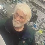 Surveillance image of an older man with white hair and a full white beard, wearing a dark green and black short-sleeved shirt, standing inside a store aisle with bicycles visible in the background.