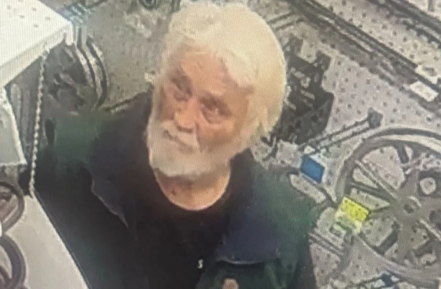 Surveillance image of an older man with white hair and a full white beard, wearing a dark green and black short-sleeved shirt, standing inside a store aisle with bicycles visible in the background.