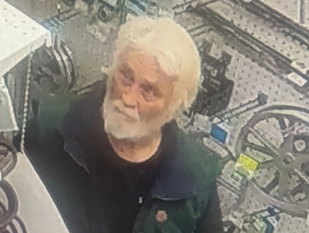 Surveillance image of an older man with white hair and a full white beard, wearing a dark green and black short-sleeved shirt, standing inside a store aisle with bicycles visible in the background.