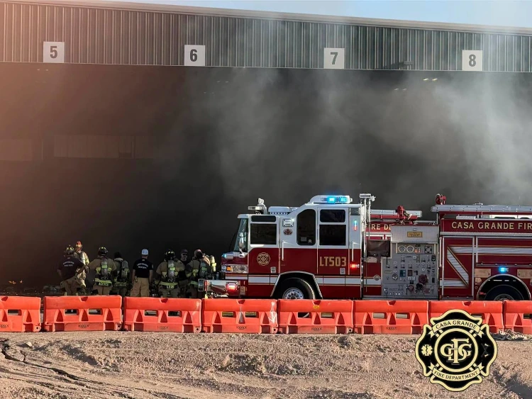 Casa Grande firefighters stand behind orange traffic barriers beside a ladder truck as smoke drifts from the transfer stations numbered loading bays