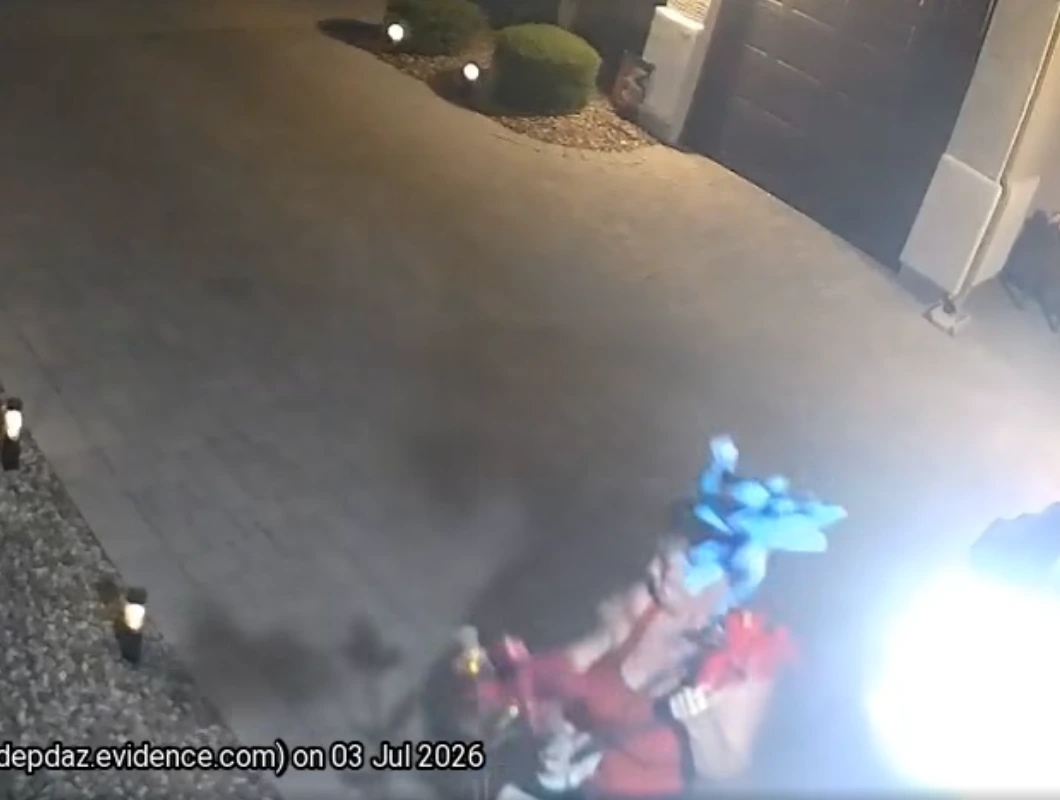 Overhead nighttime surveillance image of a residential driveway with landscape lighting, showing a person near the bottom of the frame carrying blue and red yard decorations