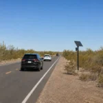 "A solar-powered Flock license plate camera on a pole beside a two-lane desert road in Pinal County, Arizona, with vehicles passing by.