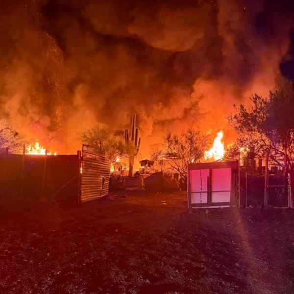 Flames and heavy smoke rise from a mobile home and outbuildings at night during the Apache Junction mobile home fire, with saguaro cactus silhouetted against the blaze.