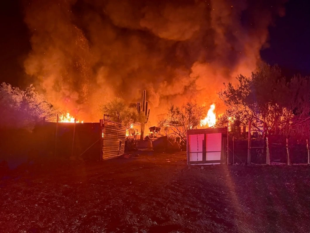 Flames and heavy smoke rise from a mobile home and outbuildings at night during the Apache Junction mobile home fire, with saguaro cactus silhouetted against the blaze.