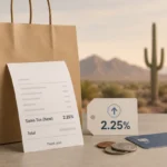 A paper shopping bag, receipt reading “Sales Tax (New) 2.25%,” a 2.25% tag, coins, and a payment card sit on a tabletop against a blurred Sonoran desert backdrop with mountains and a saguaro cactus, illustrating San Tan Valley’s new town sales tax.