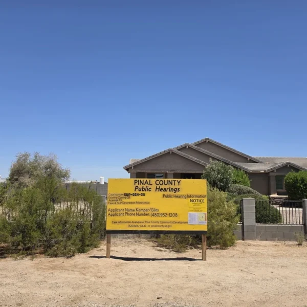 A large yellow "PINAL COUNTY Public Hearings" notice sign stands in the dirt in front of a single-story tan stucco home with a low block wall and wrought-iron fencing. The sign lists case number SUP-024-25, an existing zoning of GR, an acreage of 1.5, a case description of "Microschool," an applicant name of Kemper/Giles, and an applicant phone number, along with a smaller public hearing notice attached to its face. The setting is a rural desert lot under a clear blue sky.
