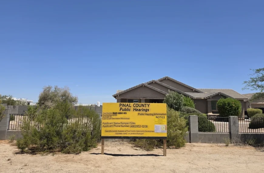 A large yellow "PINAL COUNTY Public Hearings" notice sign stands in the dirt in front of a single-story tan stucco home with a low block wall and wrought-iron fencing. The sign lists case number SUP-024-25, an existing zoning of GR, an acreage of 1.5, a case description of "Microschool," an applicant name of Kemper/Giles, and an applicant phone number, along with a smaller public hearing notice attached to its face. The setting is a rural desert lot under a clear blue sky.
