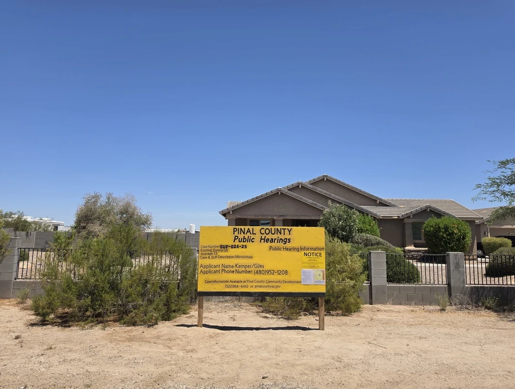 A large yellow "PINAL COUNTY Public Hearings" notice sign stands in the dirt in front of a single-story tan stucco home with a low block wall and wrought-iron fencing. The sign lists case number SUP-024-25, an existing zoning of GR, an acreage of 1.5, a case description of "Microschool," an applicant name of Kemper/Giles, and an applicant phone number, along with a smaller public hearing notice attached to its face. The setting is a rural desert lot under a clear blue sky.