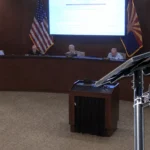 A Flock Safety license plate reader camera on a solar-powered pole appears in the foreground while four Pinal County supervisors sit at the dais during the Aug. 12 meeting.