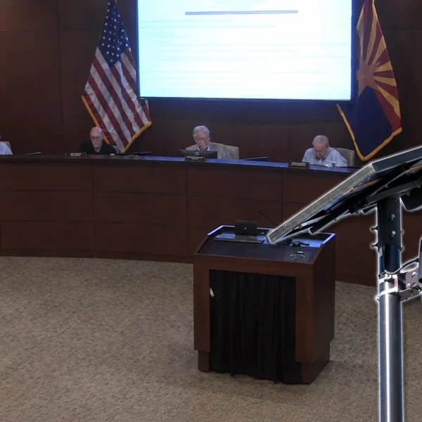 A Flock Safety license plate reader camera on a solar-powered pole appears in the foreground while four Pinal County supervisors sit at the dais during the Aug. 12 meeting.