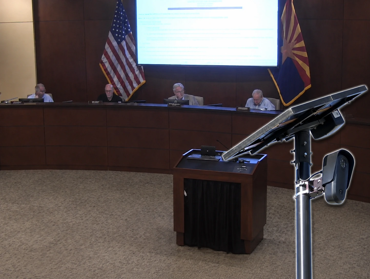 A Flock Safety license plate reader camera on a solar-powered pole appears in the foreground while four Pinal County supervisors sit at the dais during the Aug. 12 meeting.