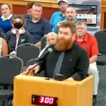 Adam Power, a bearded man in a dark shirt, speaks at a wooden podium with a microphone during the Casa Grande City Council public comment period, with a three-minute timer showing 3:00 and audience members seated behind him.