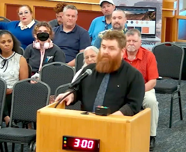 Adam Power, a bearded man in a dark shirt, speaks at a wooden podium with a microphone during the Casa Grande City Council public comment period, with a three-minute timer showing 3:00 and audience members seated behind him.