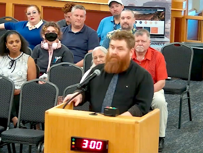 Adam Power, a bearded man in a dark shirt, speaks at a wooden podium with a microphone during the Casa Grande City Council public comment period, with a three-minute timer showing 3:00 and audience members seated behind him.