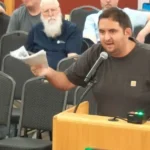A resident speaks at a podium during a city council meeting, holding up a stack of printed pages.