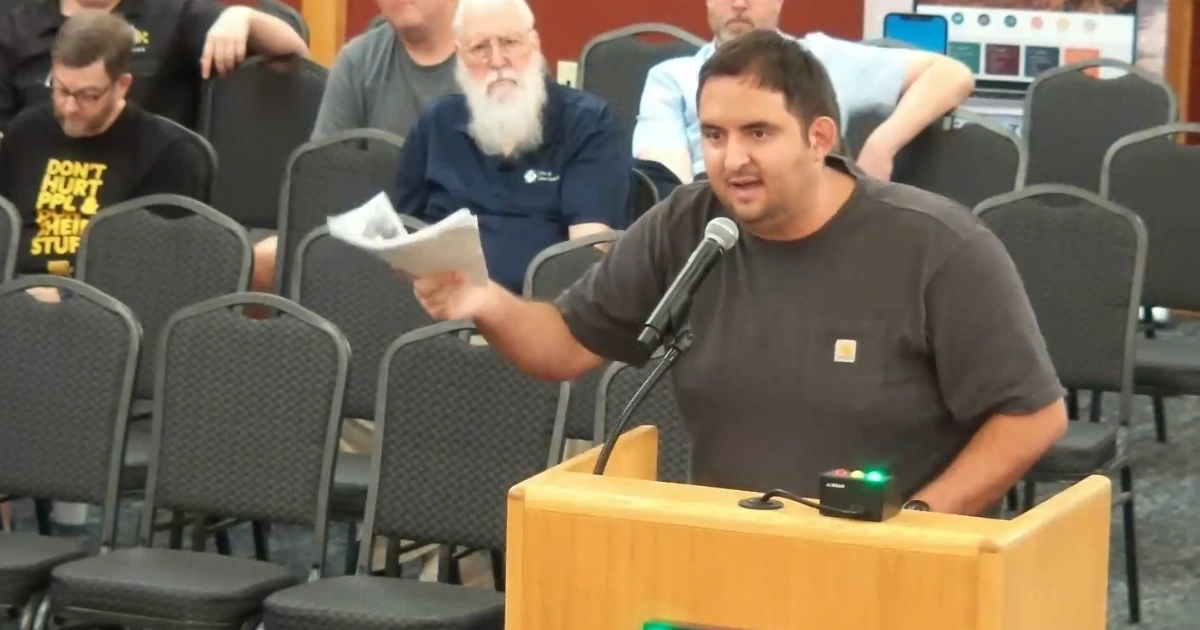 A resident speaks at a podium during a city council meeting, holding up a stack of printed pages.