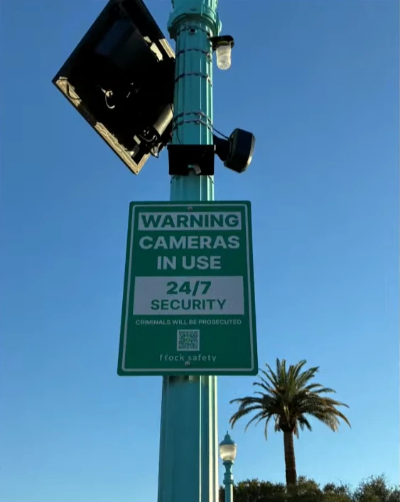 A solar-powered Flock community video camera mounted above a green 24-hour surveillance warning sign.