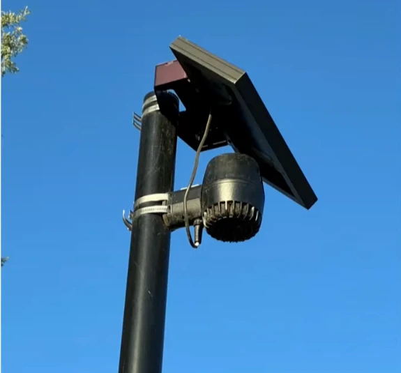 A solar-powered gunshot detection sensor mounted on a pole against a blue sky.